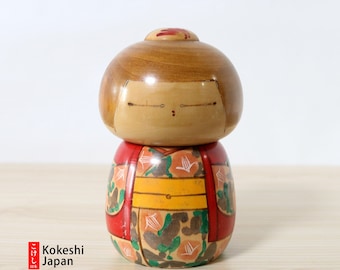May include: A traditional Japanese Kokeshi doll with a red, yellow, and green patterned kimono. The doll has a round wooden body and a painted face.