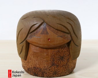 May include: A traditional Japanese Kokeshi doll carved from wood. The doll has a round body with a carved floral pattern and a simple face with a red dot for a mouth. The doll's hair is carved in a flowing style.