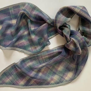 May include: A patterned scarf in blue, green, and purple with a grey border. The scarf is knotted. The scarf is made of a lightweight material. A small square tag is on one end.