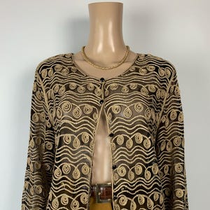 May include: A sheer, long-sleeved, open-front jacket with a gold swirling pattern over a black base. The jacket has a round neckline and is buttoned with black buttons. A gold necklace and brown belt are also visible.