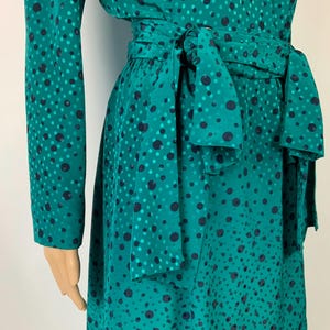 May include: A teal dress with a polka dot design. The long-sleeved dress has a matching tie belt at the waist. The polka dots are in shades of teal and navy blue. The dress is made of a silky material.
