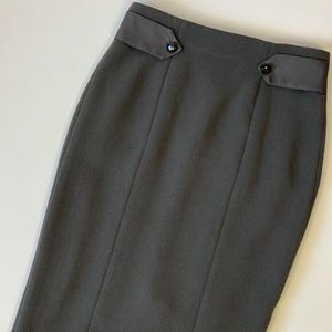 May include: A charcoal gray pencil skirt with a tailored design. The skirt features a high waist with two decorative dark gray fabric panels, each adorned with a small, round, black button. Vertical seams run down the front, adding structure to the garment.