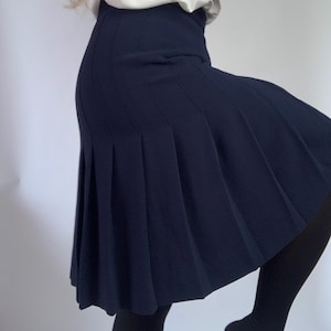 Può includere: Gonna plissettata blu navy. La gonna ha una vita alta e arriva alle ginocchia. Il tessuto sembra essere un materiale liscio e tessuto. La gonna è abbinata a una camicetta bianca.