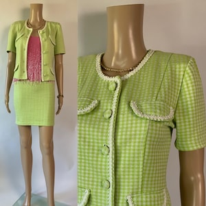 May include: A lime green and white checked skirt suit on a mannequin. The suit features a short-sleeved jacket with white trim and pockets, and a matching mini skirt. A pink top with fringe is visible. A gold necklace is also visible.