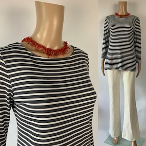 Puede incluir: Un maniquí exhibe una camiseta a rayas azul marino y blanca con pantalones color crema. Un collar de coral rojo adorna el cuello. El conjunto se presenta sobre un fondo blanco. La camiseta es de manga larga.