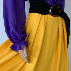 May include: A striking vintage dress in a colour block design. The top is purple with long sleeves and a textured pattern, and the skirt is bright yellow. A black belt with a large brown buckle defines the waist, creating a bold silhouette.