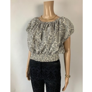May include: A cream-colored, short-sleeved top with a black geometric pattern. The top has a gathered neckline and a smocked waist. The model is also wearing black patterned pants and a gold necklace and bracelet.