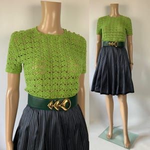 May include: A lime green crochet top with short sleeves and gold shoulder buttons. The top is paired with a dark green belt with a gold buckle and a grey and black striped skirt. The outfit is displayed on a mannequin.