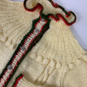 May include: Cream-colored knitted cardigan with red and green trim. The cardigan features decorative silver buttons and a ruffled collar. The sleeves and front are adorned with textured details, creating a cozy, handcrafted look.