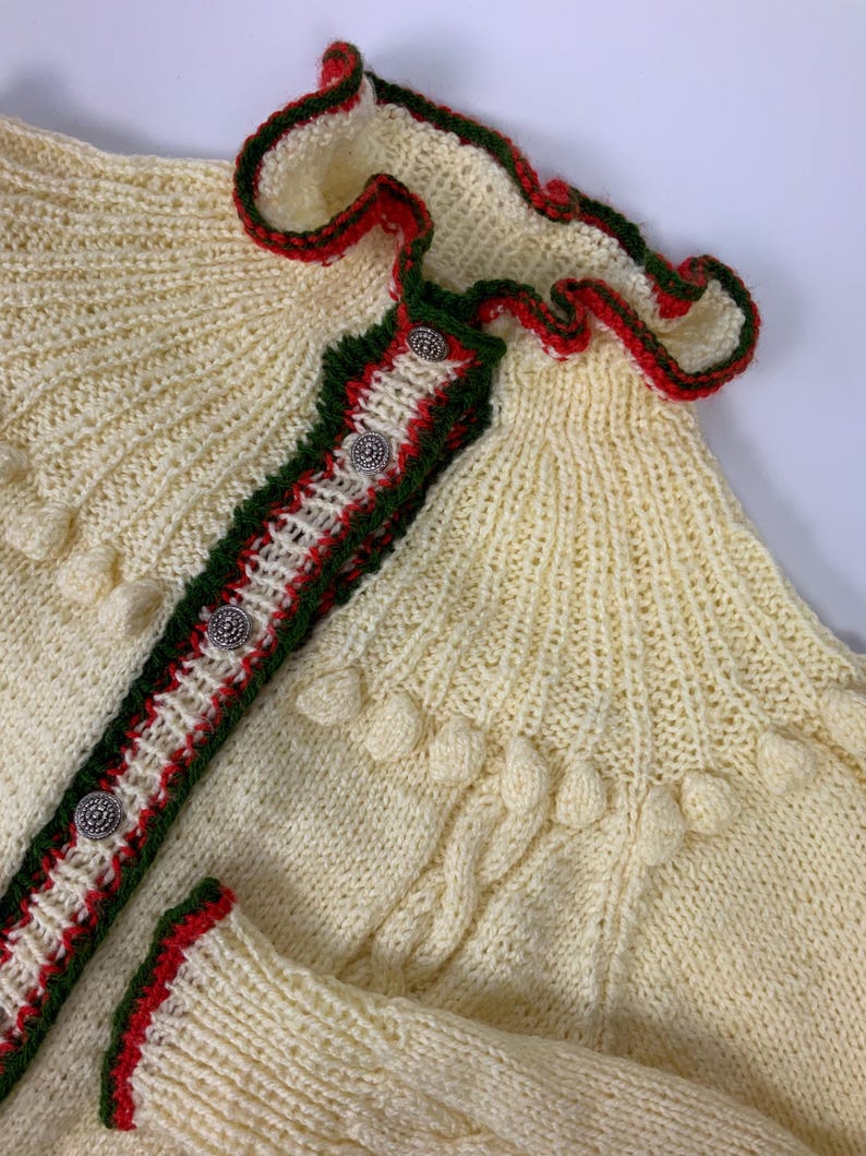 May include: Cream-colored knitted cardigan with red and green trim. The cardigan features decorative silver buttons and a ruffled collar. The sleeves and front are adorned with textured details, creating a cozy, handcrafted look.