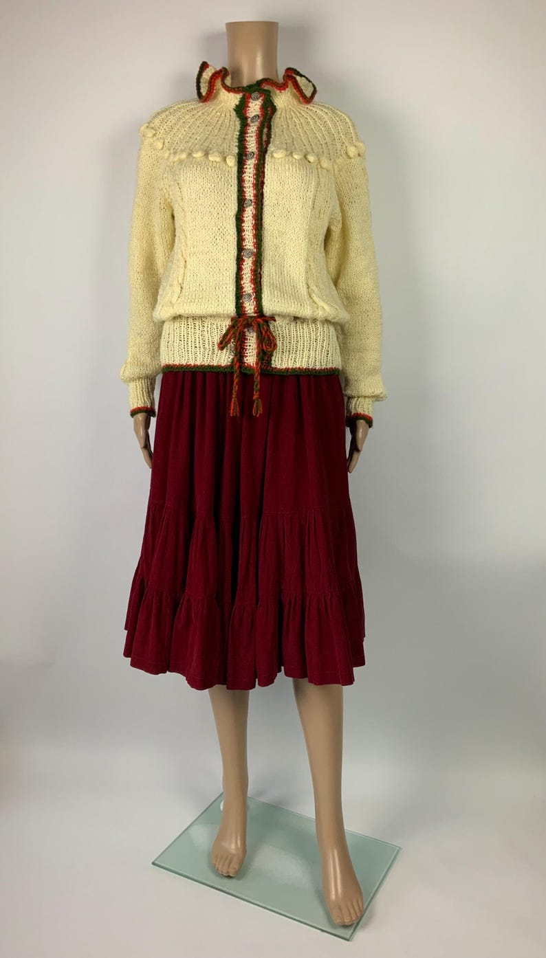 May include: A vintage cream-colored knit sweater with a decorative collar and trim, paired with a tiered burgundy skirt. The sweater has a button-up front with green and red accents. The skirt is a midi length.