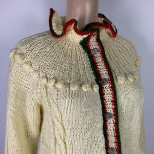 May include: Cream-colored knitted cardigan with a ruffled collar trimmed in red and green. The front features a red and green stripe with decorative buttons. The sleeves have a cable knit design and the collar has a decorative trim.