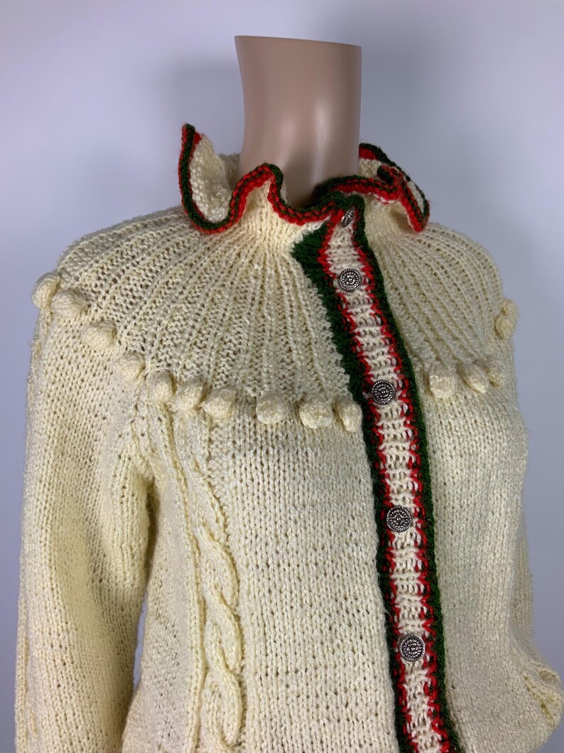 May include: Cream-colored knitted cardigan with a ruffled collar trimmed in red and green. The front features a red and green stripe with decorative buttons. The sleeves have a cable knit design and the collar has a decorative trim.