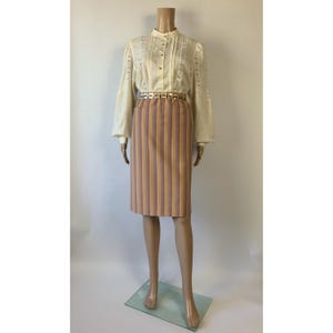 Op de afbeelding: Een crèmekleurige blouse met lange mouwen, verticale kanten inzetstukken en een knoopsluiting, gecombineerd met een knielange rok met verticale strepen in roze, geel en bruin, geaccentueerd door een metallic goudkleurige tailleceintuur.