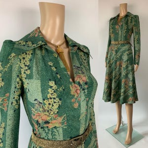 May include: Green vintage dress with a floral pattern, long sleeves, and a collared neckline. The dress features a gold-colored belt and a gold necklace. The dress is displayed on a mannequin.