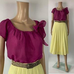 以下が含まれることがあります： マゼンタ色のオフショルダーのトップスとイエローのプリーツスカートのツーピースの衣装。リボンディテールとゴールドカラーのベルトがアクセント。マネキンに着せられた状態で展示されています。