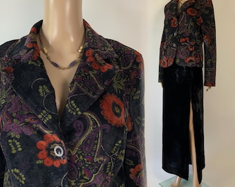 May include: A long-sleeve blazer featuring a dark gray base with a vibrant floral and paisley pattern in shades of orange, purple, and green, paired with a solid black floor-length skirt that includes a high side slit and a gold-toned necklace.