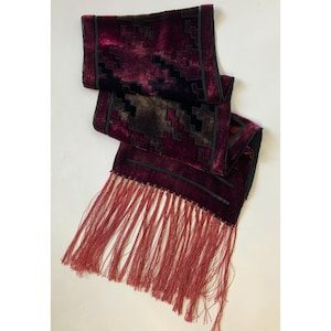 May include: A deep burgundy velvet scarf featuring a dark geometric pattern and a black border. The scarf is finished with long, thin, matching burgundy fringe tassels along the bottom edge, creating a textured and layered appearance.