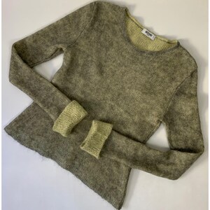 May include: An olive green long-sleeved sweater with light green cuffs and neckline. The sweater has a textured appearance. The label "MOSCHINO" is visible on the neckline. The cuffs are a lighter shade of green.