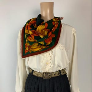 Puede incluir: Un pañuelo cuadrado con fondo verde oscuro, grandes motivos florales naranjas y amarillos, y un borde color óxido con bordes deshilachados. El pañuelo se lleva sobre una blusa color crema de manga larga con detalles de encaje y un cinturón dorado.