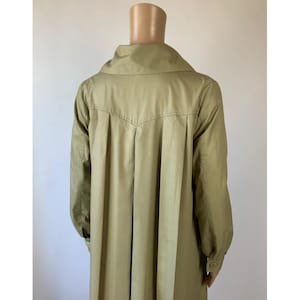 May include: A light olive green trench coat featuring a wide collar, long sleeves with buttoned cuffs, and a back yoke with vertical pleats that extend down the length of the garment. The coat is displayed on a mannequin against a plain background.