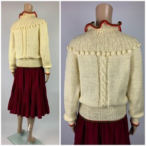 May include: Cream-colored knit sweater with a high collar and bobble trim. The sweater features a cable knit design down the back and is paired with a tiered burgundy skirt. Red and green trim accents the collar, cuffs, and skirt hem.
