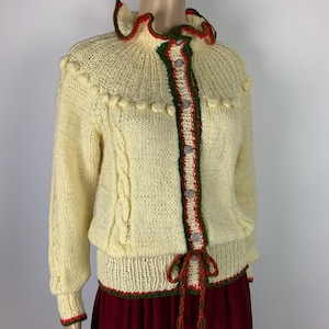 May include: Cream-colored knitted cardigan with a ruffled collar and red and green trim. The sweater features a cable knit design on the sleeves and decorative bobbles. It has a button-up front and a tie closure at the waist, offering a vintage aesthetic.