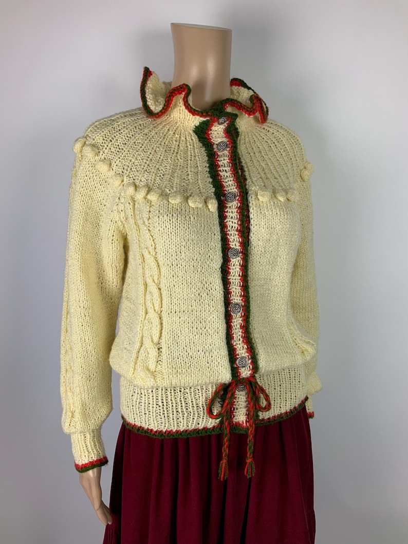 May include: Cream-colored knitted cardigan with a ruffled collar and red and green trim. The sweater features a cable knit design on the sleeves and decorative bobbles. It has a button-up front and a tie closure at the waist, offering a vintage aesthetic.