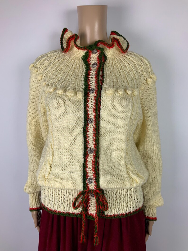May include: Cream-colored knitted cardigan with a ruffled collar and decorative pom-poms. The trim and button placket feature red and green accents. The cardigan has a tie closure at the waist and is displayed on a mannequin.