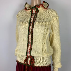 May include: Cream-colored knitted cardigan with red and green trim. The sweater features a ruffled collar, decorative buttons, and a tie closure. The sleeves have a slight ruffle at the wrist. The cardigan is paired with a burgundy skirt.