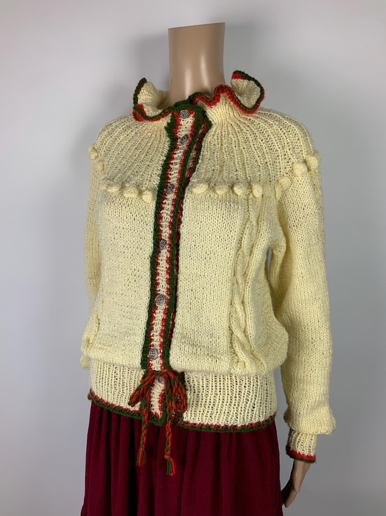 May include: Cream-colored knitted cardigan with red and green trim. The sweater features a ruffled collar, decorative buttons, and a tie closure. The sleeves have a slight ruffle at the wrist. The cardigan is paired with a burgundy skirt.