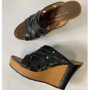 May include: Black leather wedge sandals with a woven strap design. The sandals have a wooden platform and wedge heel. The brand name "pierre cardin" is visible on the insole. The sandals are a size 8.
