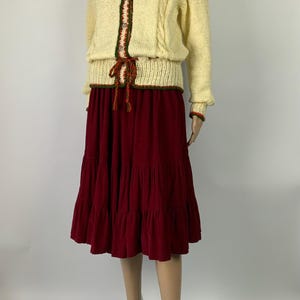 May include: A two-piece outfit featuring a cream-colored knit sweater with a decorative collar and trim, paired with a tiered burgundy skirt. The sweater has a decorative trim with green and red accents. The skirt is a deep burgundy color.
