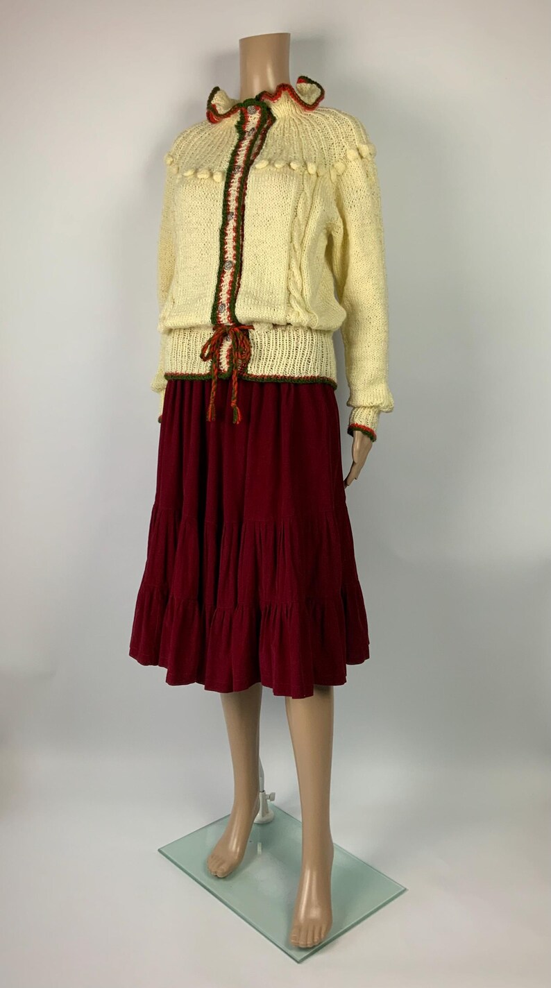 May include: A two-piece outfit featuring a cream-colored knit sweater with a decorative collar and trim, paired with a tiered burgundy skirt. The sweater has a decorative trim with green and red accents. The skirt is a deep burgundy color.