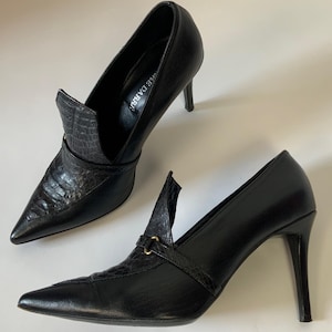 May include: A pair of black leather high heels with pointed toes. The shoes have a textured, reptile-like design on the vamps and straps, with a small gold-coloured buckle. The heels are stiletto style.