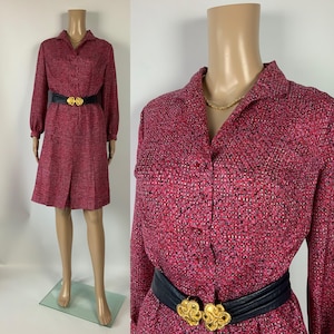 Puede incluir: Un vestido vintage con estampado rojo y negro con mangas largas y cuello camisero. El vestido presenta un cinturón negro con una hebilla dorada. El vestido se exhibe en un maniquí.