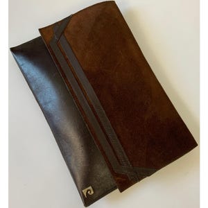 May include: Two leather wallets. One is a dark brown, smooth leather with a gold emblem. The other is a brown suede with dark brown leather trim. Both wallets are rectangular and appear to be long.