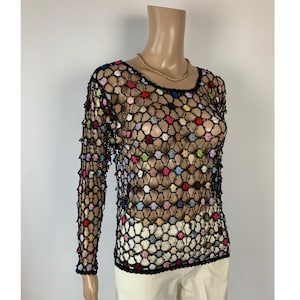 Peut inclure: Un haut à manches longues présentant un motif en filet crocheté noir entremêlé de petits motifs floraux circulaires multicolores dans des tons de rouge, bleu, jaune, rose et blanc. Le vêtement possède une encolure ronde et une structure semi-transparente.