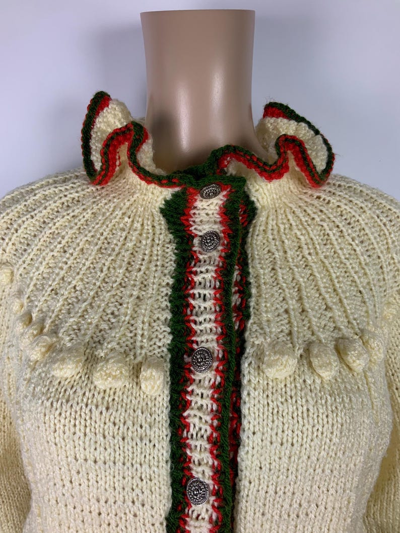 May include: Cream-colored knitted cardigan with a ruffled collar and trim in green and red. The cardigan features decorative buttons and textured knit detailing. The garment is displayed on a mannequin.