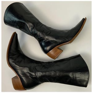 Peut inclure: Paire de bottes en cuir noir avec bouts pointus et talons en bois. Les bottes ont un design classique, avec une hauteur mi-mollet et une finition élégante et polie. Les talons mesurent environ 5 cm de haut.