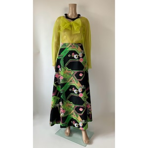 Op de afbeelding: Een blouse met lange mouwen van doorschijnende chartreuse stof met een grote strik bij de halslijn, gecombineerd met een zwarte rok tot op de grond met een geometrisch patroon van groene lijnen en roze en witte bloemmotieven.