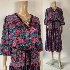 May include: A midi-length dress featuring a vibrant floral pattern in shades of pink, blue, and purple. The garment has a V-neckline with black polka-dot trim, three-quarter length sleeves, a smocked waist, and a thin black rope belt with gold accents.