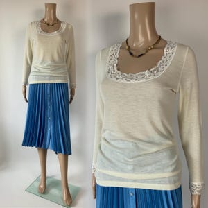 May include: A long-sleeved cream-colored top featuring a scoop neckline trimmed with white lace and matching lace cuffs, paired with a bright blue pleated midi skirt that has a vertical row of buttons down the front center.