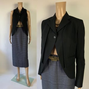 May include: A black blazer and vest set paired with a gray and black plaid pencil skirt. The blazer has a single-button closure and a unique design. The skirt is knee-length. A gold-colored belt adds a touch of elegance to the outfit. The outfit is displayed on a mannequin.