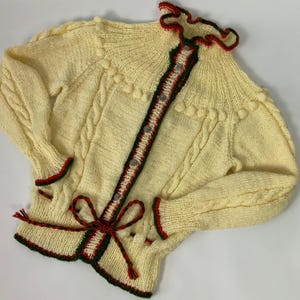 May include: A cream-colored, hand-knitted cardigan with a cable knit design. The sweater features red and green trim, decorative buttons, and a bow at the waist. The collar and cuffs have a ruffled edge. The sleeves are long.