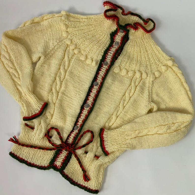 May include: A cream-colored, hand-knitted cardigan with a cable knit design. The sweater features red and green trim, decorative buttons, and a bow at the waist. The collar and cuffs have a ruffled edge. The sleeves are long.