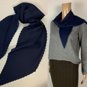 Può includere: Sciarpa plissettata blu navy con un design strutturato e ondulato. La sciarpa è esposta in due modi: drappeggiata e stilizzata attorno al collo di un manichino. I bordi della sciarpa hanno un bordo decorativo smerlato.