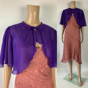 Peut inclure: Un boléro violet transparent à manches courtes évasées et fermeture à nouer sur le devant, porté sur une robe midi sans manches rose poudré avec un motif à pois circulaires ton sur ton sur tout le tissu et un ourlet évasé.