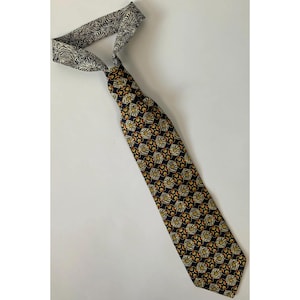 May include: A necktie featuring a dark blue base with a repeating pattern of orange and white geometric shapes. The necktie includes a contrasting neck loop with a black and white zebra-like striped pattern, visible at the top of the garment.