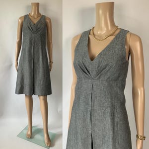 May include: A gray sleeveless dress with a V-neckline and a pleated front. The dress is displayed on a mannequin, showcasing its tailored design and knee-length hem. A gold necklace and bracelet add a touch of elegance to the outfit.